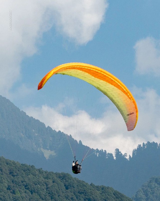 Ярко‑оранжевый параплан в небе над гористым ландшафтом, пилот в подвеске; видно обтекание крыла и общую форму крыла в потоках воздуха.