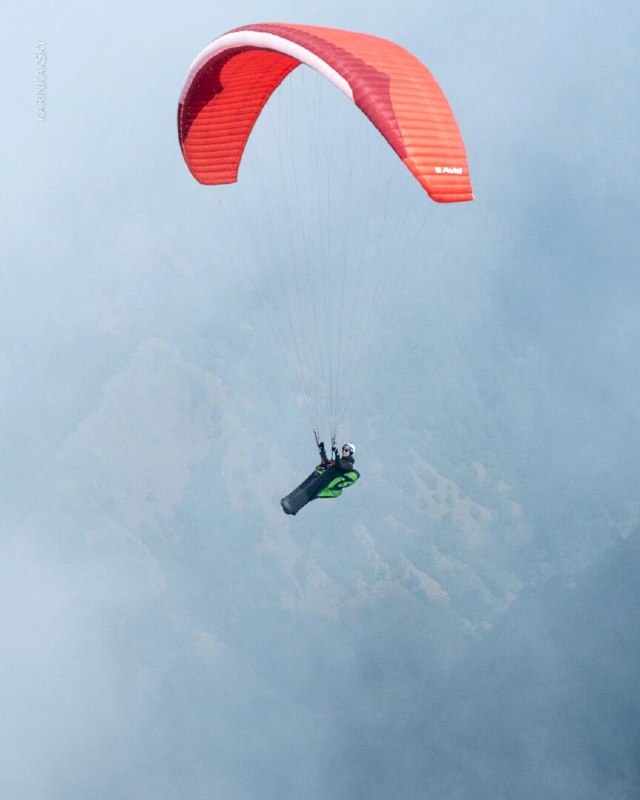 Красно‑оранжевое крыло и пилот в гамаках на фоне облачного неба; демонстрация посадочной позы, положения корпуса и натяжения строп.