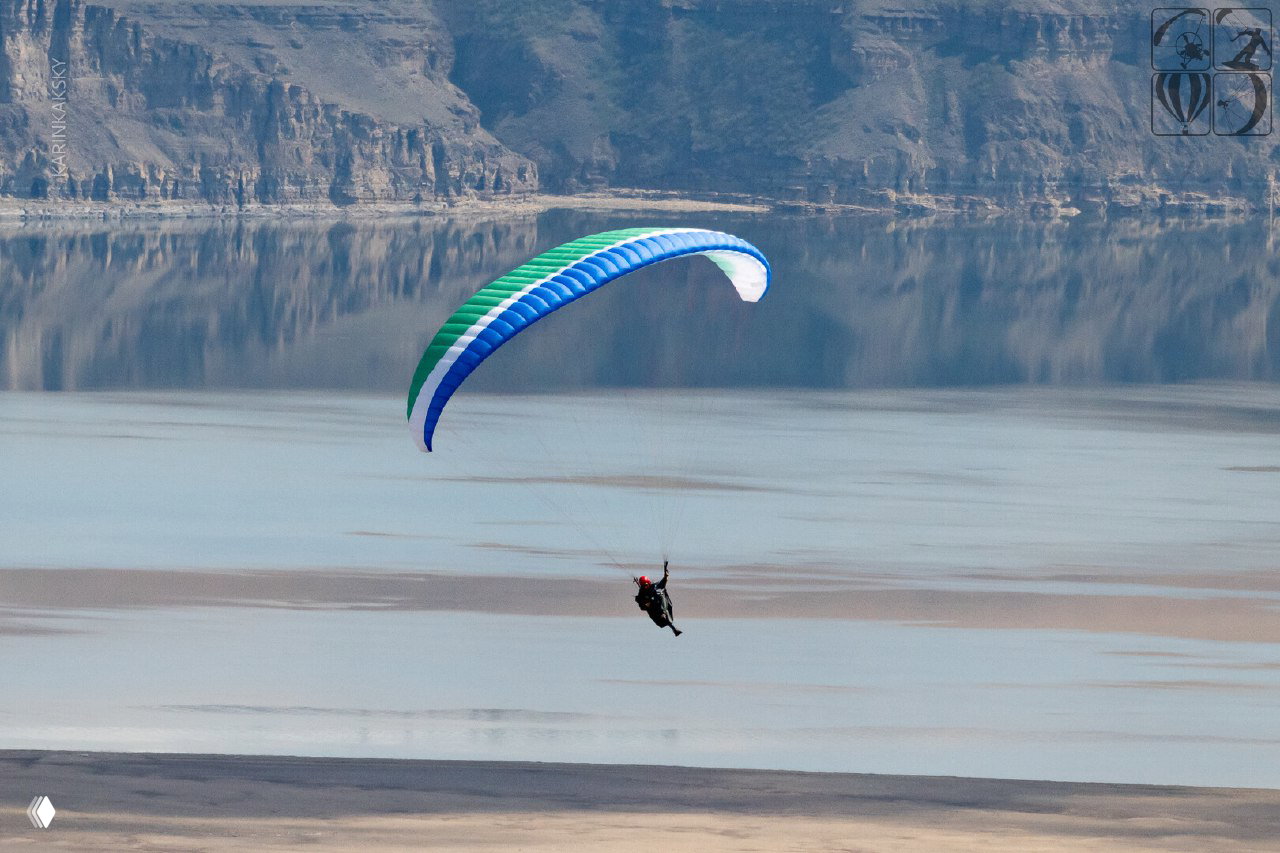 Парапланист в полёте над водной гладью и скальными берегами в зоне тренировок Хакасии — демонстрация полёта и высотной техники.