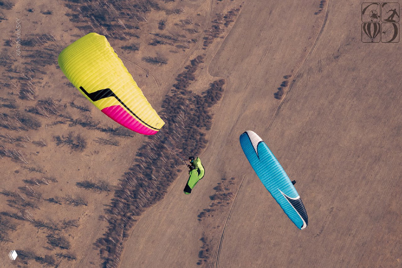 Вид сверху на несколько парапланов в небе и на земле: групповая тренировка и маршрутные полёты — кадр с аэрофотосъёмки кэмпа.