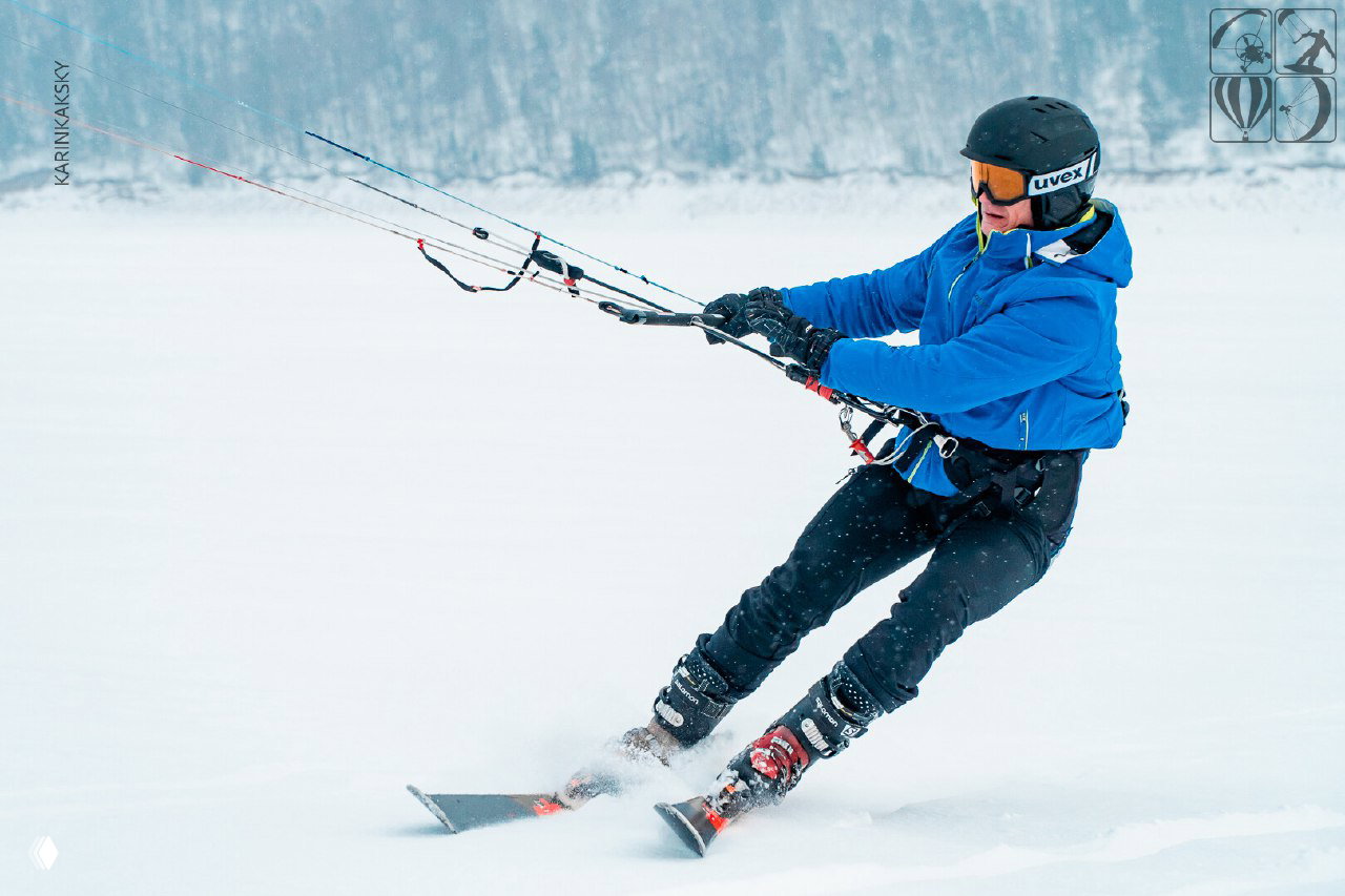 Спортсмен в синей куртке управляет кайтом на заснеженном поле; видно управление баром, следы на снегу и динамику движения.