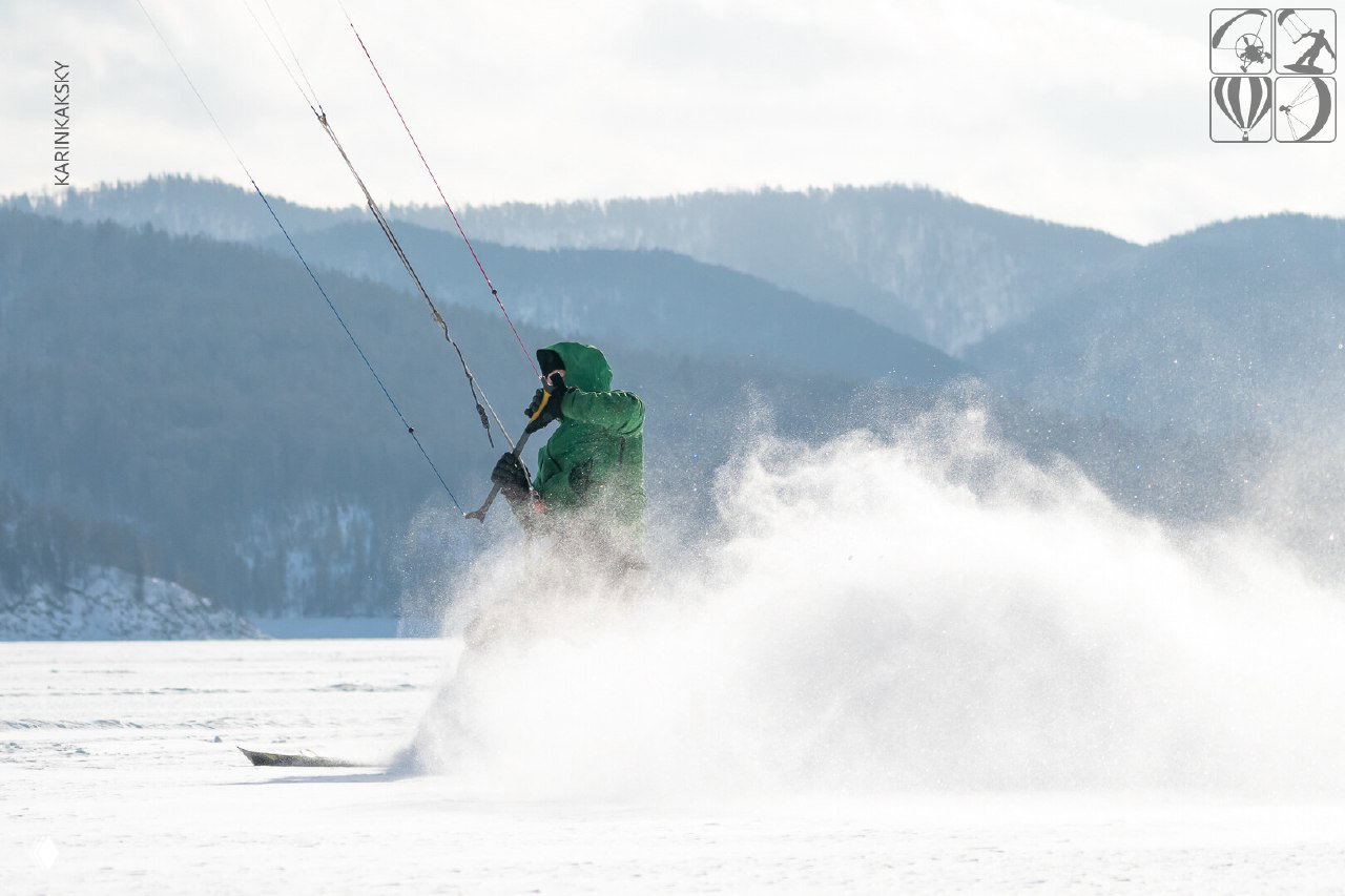Кайтер в зелёной куртке набирает скорость, снежная пыль поднимается в воздух; на фоне холмы и линия леса под облачным небом.