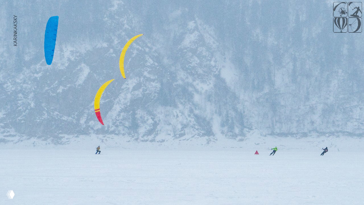 Несколько разноцветных кайтов в небе над замёрзшим озером; спортсмены распределены на дистанции по белому снежному полю.