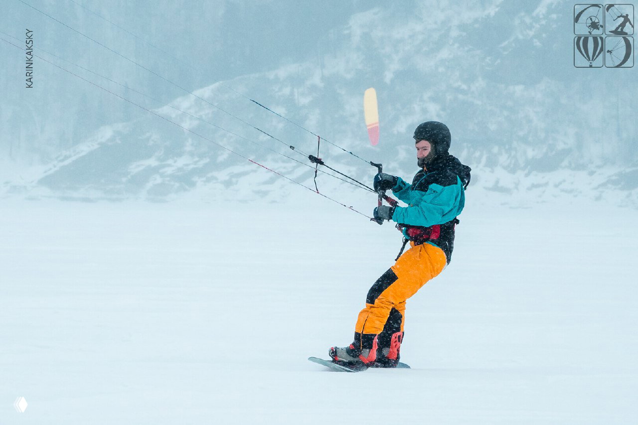 Кайтер в жёлто-оранжевых штанах едет по льду с натянутым кайтом; на заднем плане горы и слабый снегопад создают зимнюю атмосферу.