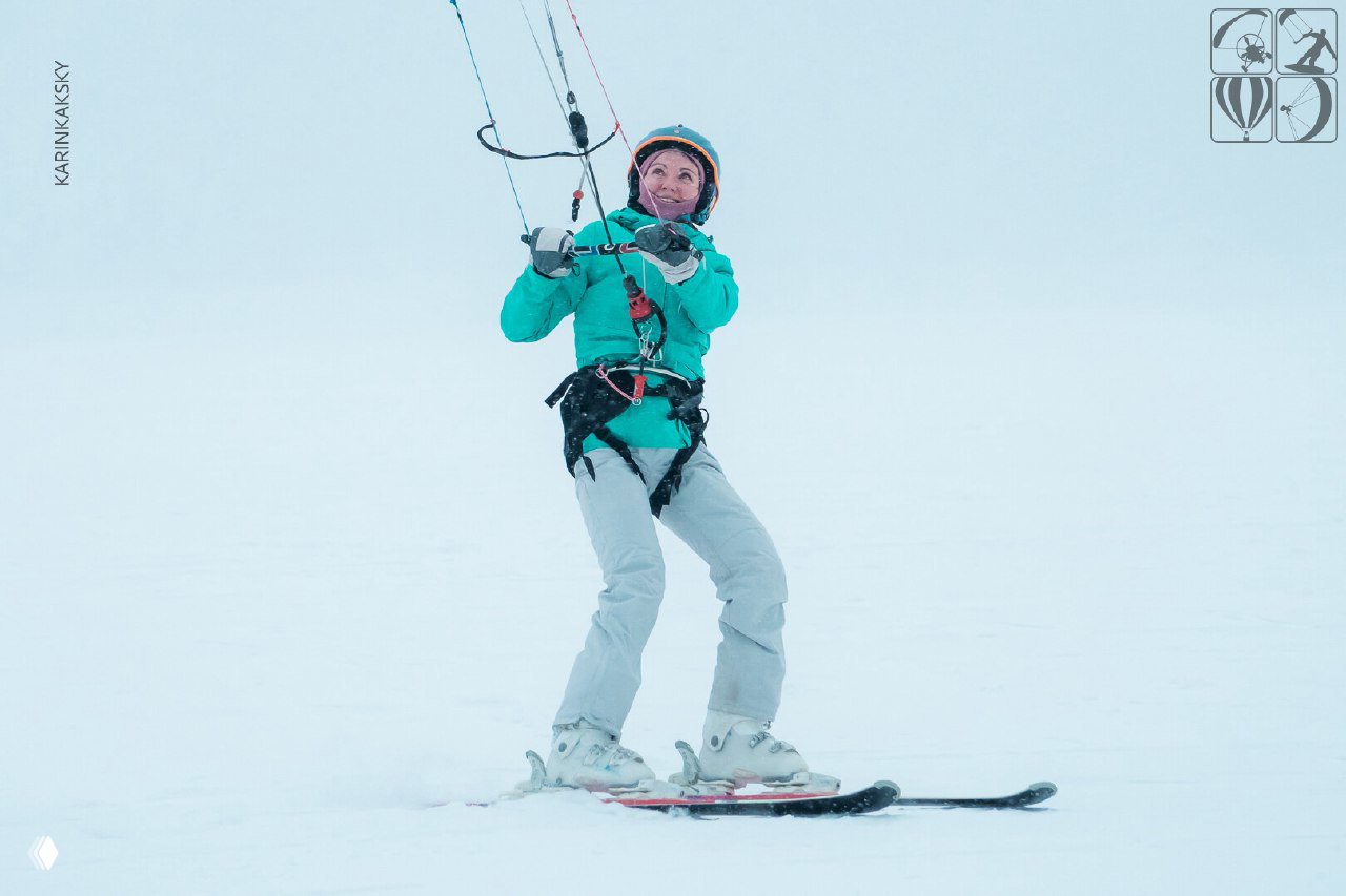 Улыбающаяся женщина на лыжах с кайтом держит бар, стоит на белом поле; кадр делает акцент на эмоциях и экипировке участника.