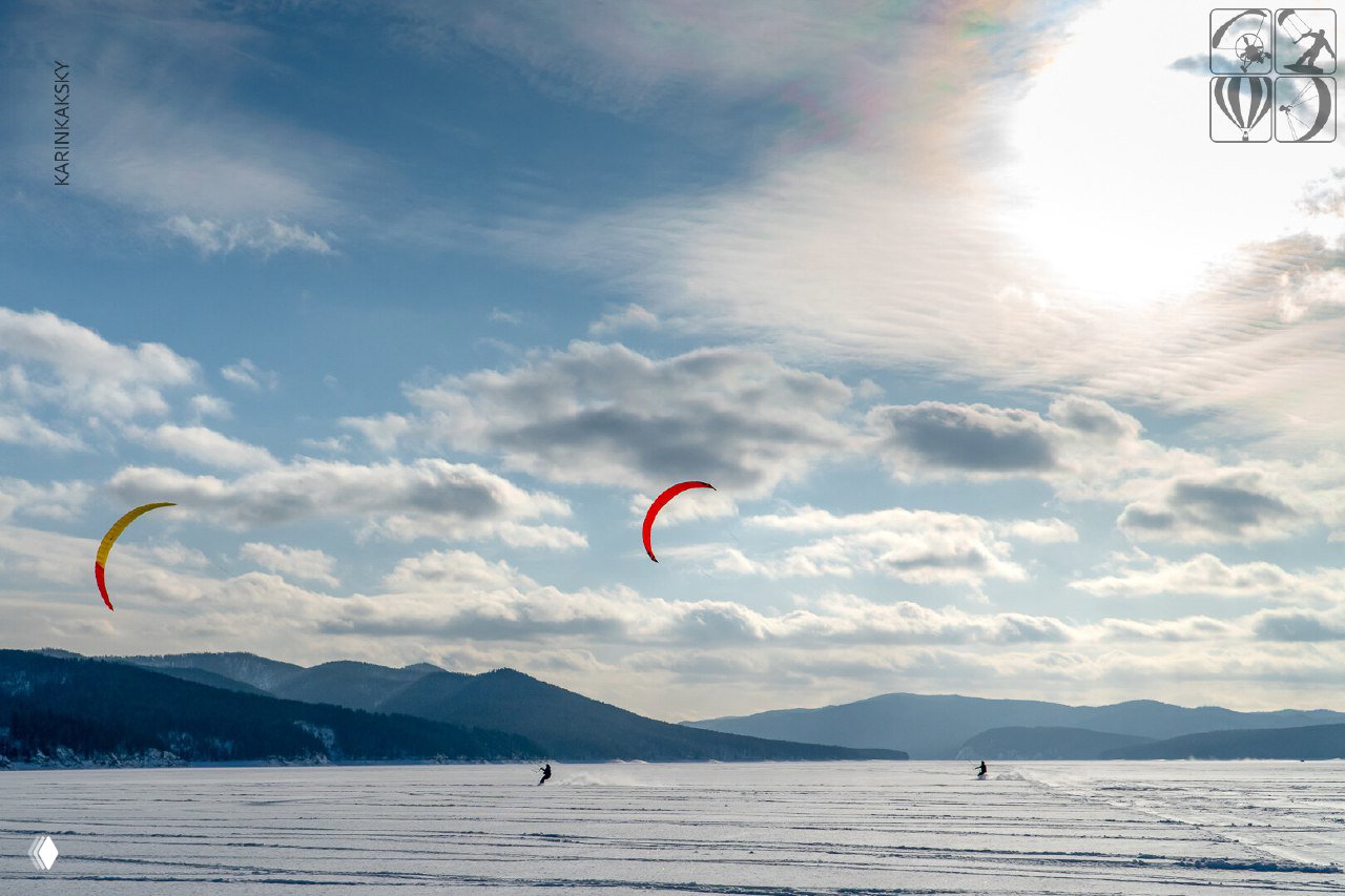 Широкий кадр с разноцветными кайтами в небе, далёкие кайтеры едут по замёрзшему озеру на фоне облачного неба и горной линии.