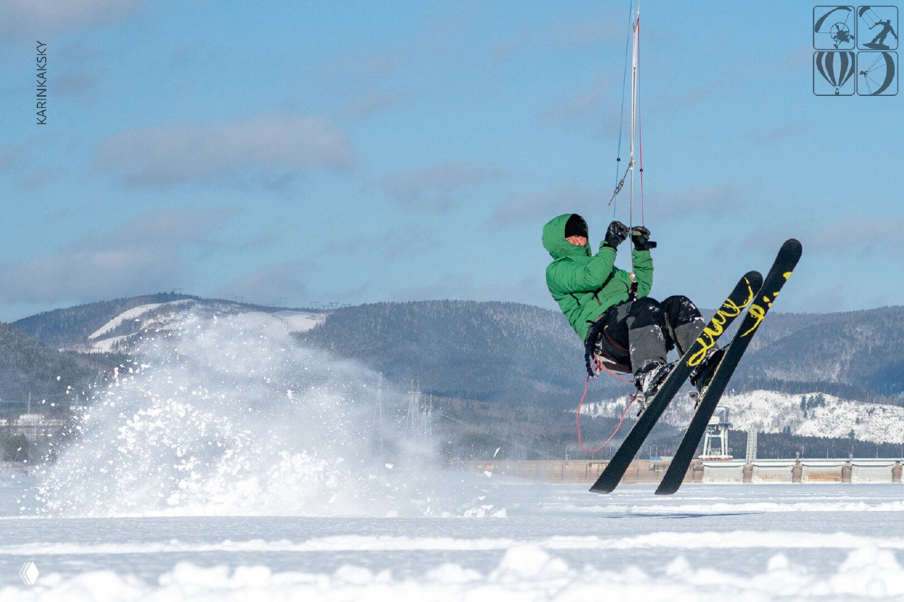 Кайтер выполняет прыжок на лыжах над снежным покровом; в кадре видны левая часть дамбы и заснеженные склоны вдали.