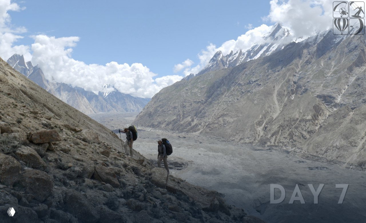 Два участника экспедиции идут с рюкзаками по каменистому склону в высокогорной долине Пакистана; на фоне видны острые пики.