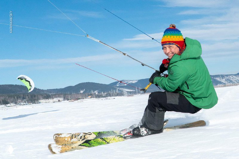 Крупный план улыбающегося спортсмена в тёплой шапке, сидящего на сноускейте с натянутыми стропами кайта; момент отдыха между заездами.