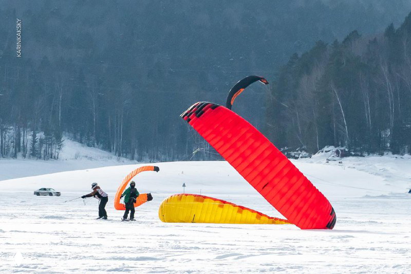 Подготовка к старту: красный купол кайта развёрнут на льду, команда проверяет стропы и оснащение перед заездом по замёрзшей поверхности.