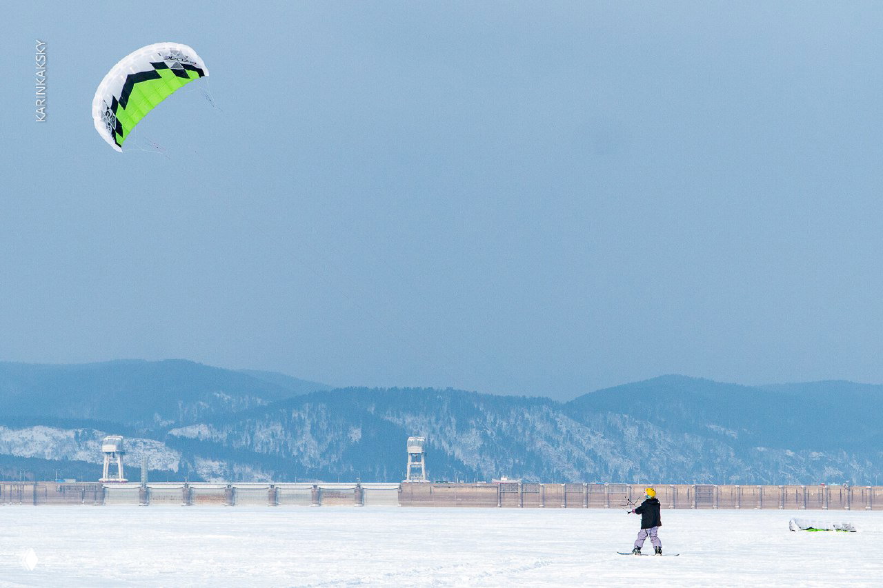 Широкий план замёрзшего озера и кайта на фоне далёких холмов и дамбы, подчёркнуты пространство и погодные условия в марте.
