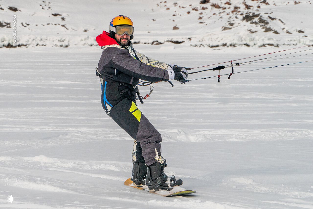 Человек в шлеме на сноуборде с кайтом, крупный план фигуры на льду, видна снаряга и динамика движения во время каталки.
