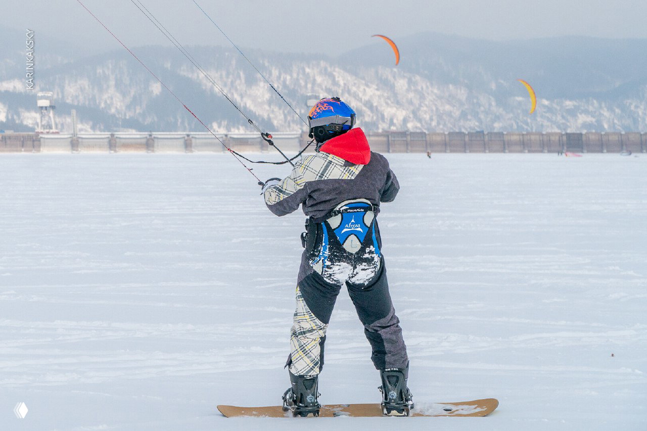Райдер на сноуборде снимается сзади: видно снаряжение, следы на снегу и несколько кайтов на горизонте — атмосфера тренировочного дня.