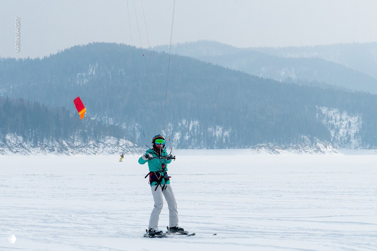 Ещё один райдер на сноуборде на фоне холмов и далёких деревень, купол кайта в небе демонстрирует масштаб пространства каталки.