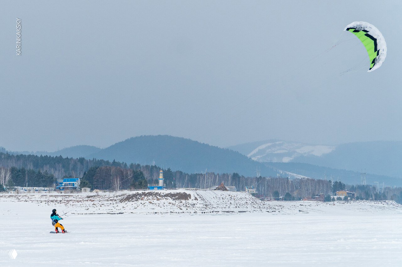 Широкий кадр: сноубордист на замёрзшем озере с крупным кайтом в небе и заснеженной прибрежной полосой, характерный мартовский свет.