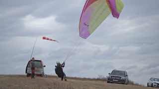 Пилот запускает параплан на ветреном поле в Хакасии: видны разноцветный купол, автомобиль поддержки, флюгер и пасмурное небо