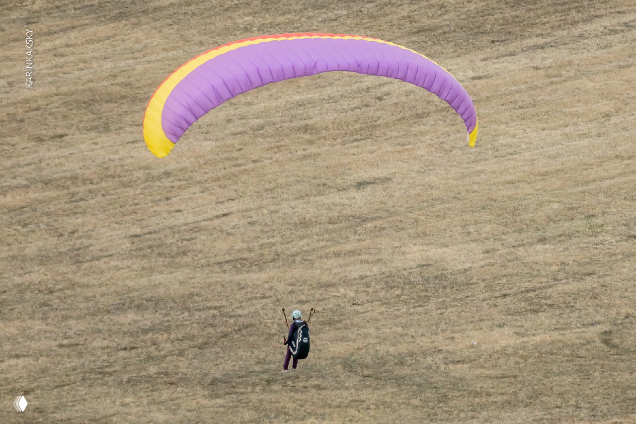 Пилот под ярко-фиолетовым куполом параплана парит над травянистым склоном — вид тренировочного полёта и освоения навыков в воздухе.