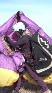 Учебная сцена: пилот в шлеме складывает фиолетовый параплан и перебирает стропы у себя на плече, подготовка к сбору и проверке снаряжения.