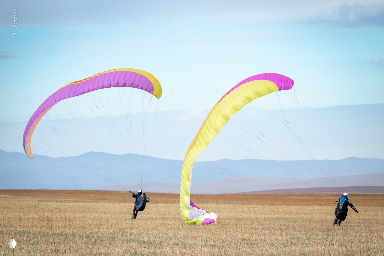 Два ученика с разноцветными крыльями поднимаются и учатся запускать парапланы на ровном поле с дальними холмами.