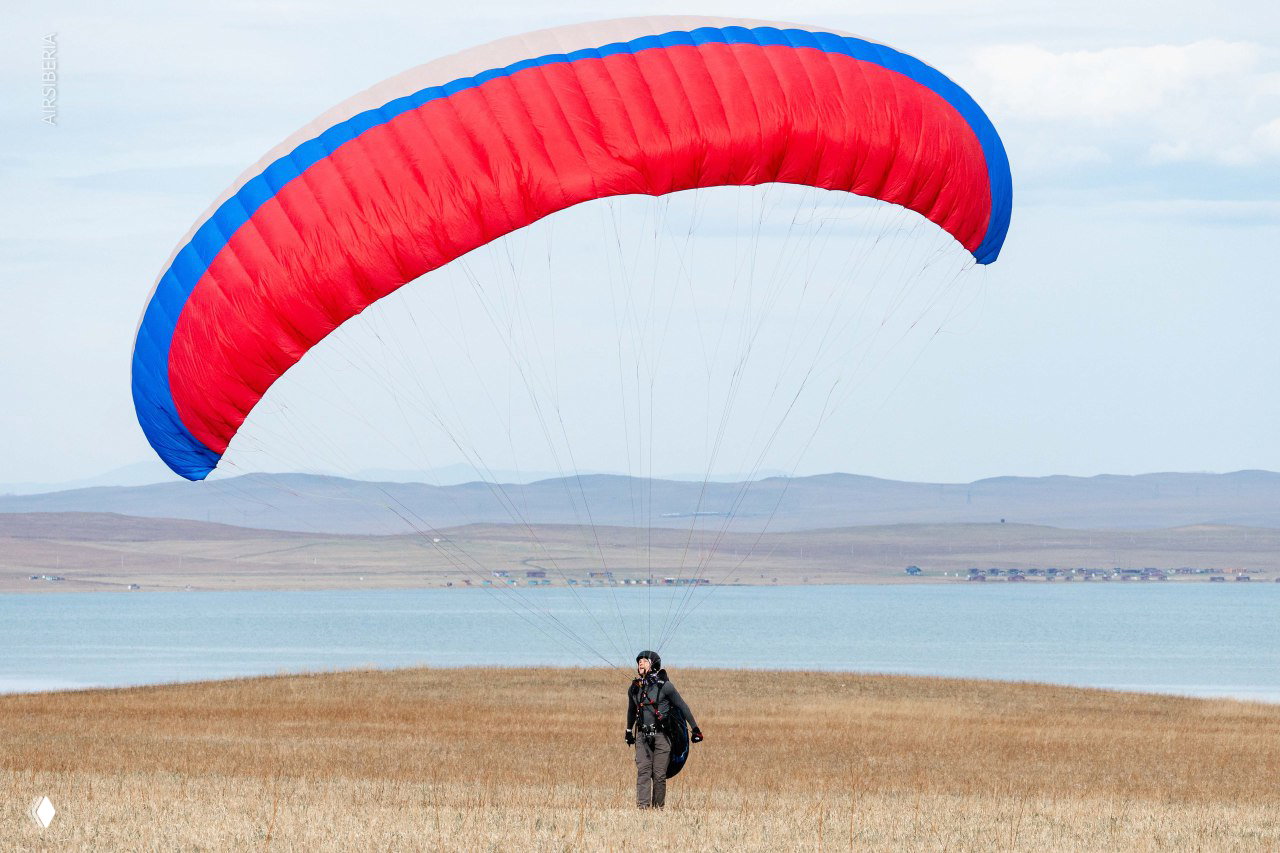 Пилот идёт по траве под полностью надутым красно-синим крылом, на заднем плане виднеется озеро и холмы.