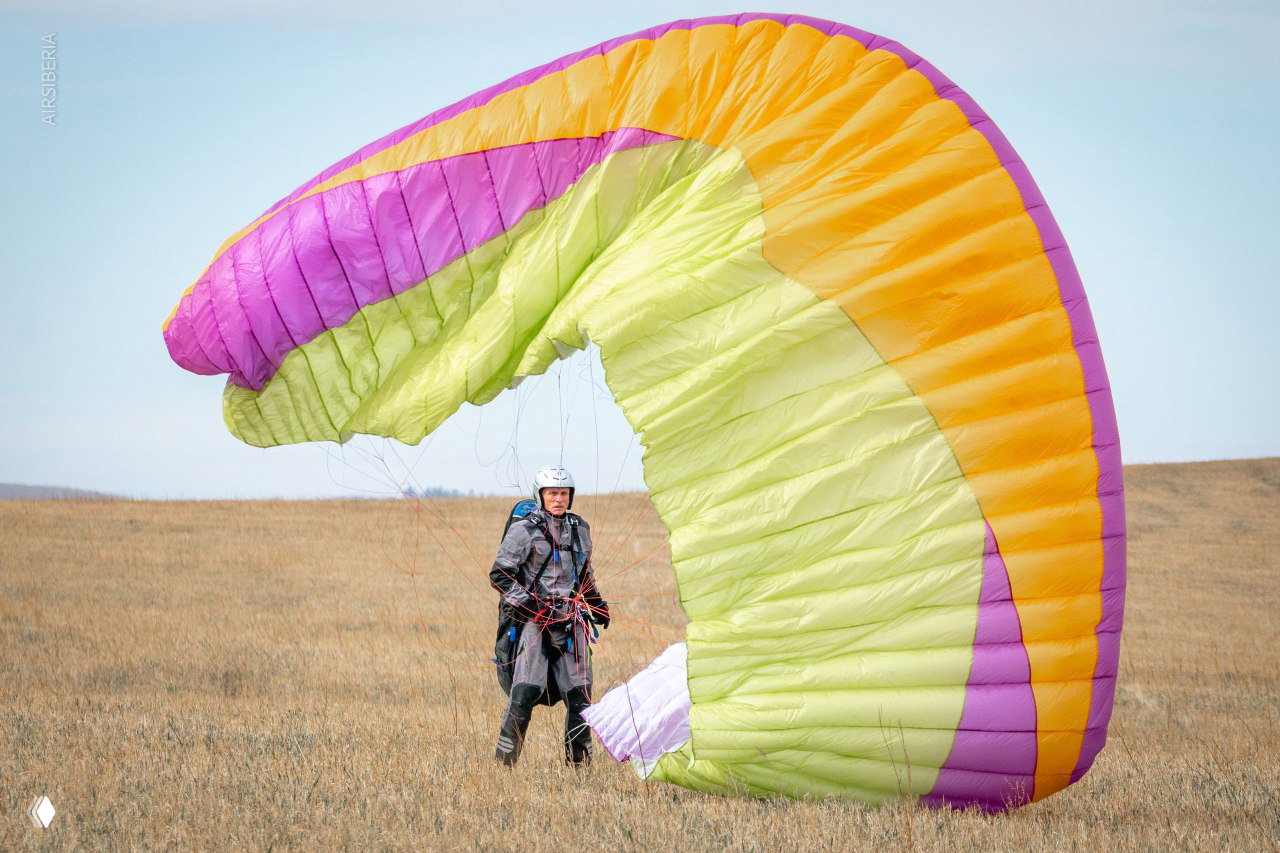 Человек в амуниции стоит рядом с ярко-зелёно-фиолетовым парапланом, который частично надут на ветру на поле.