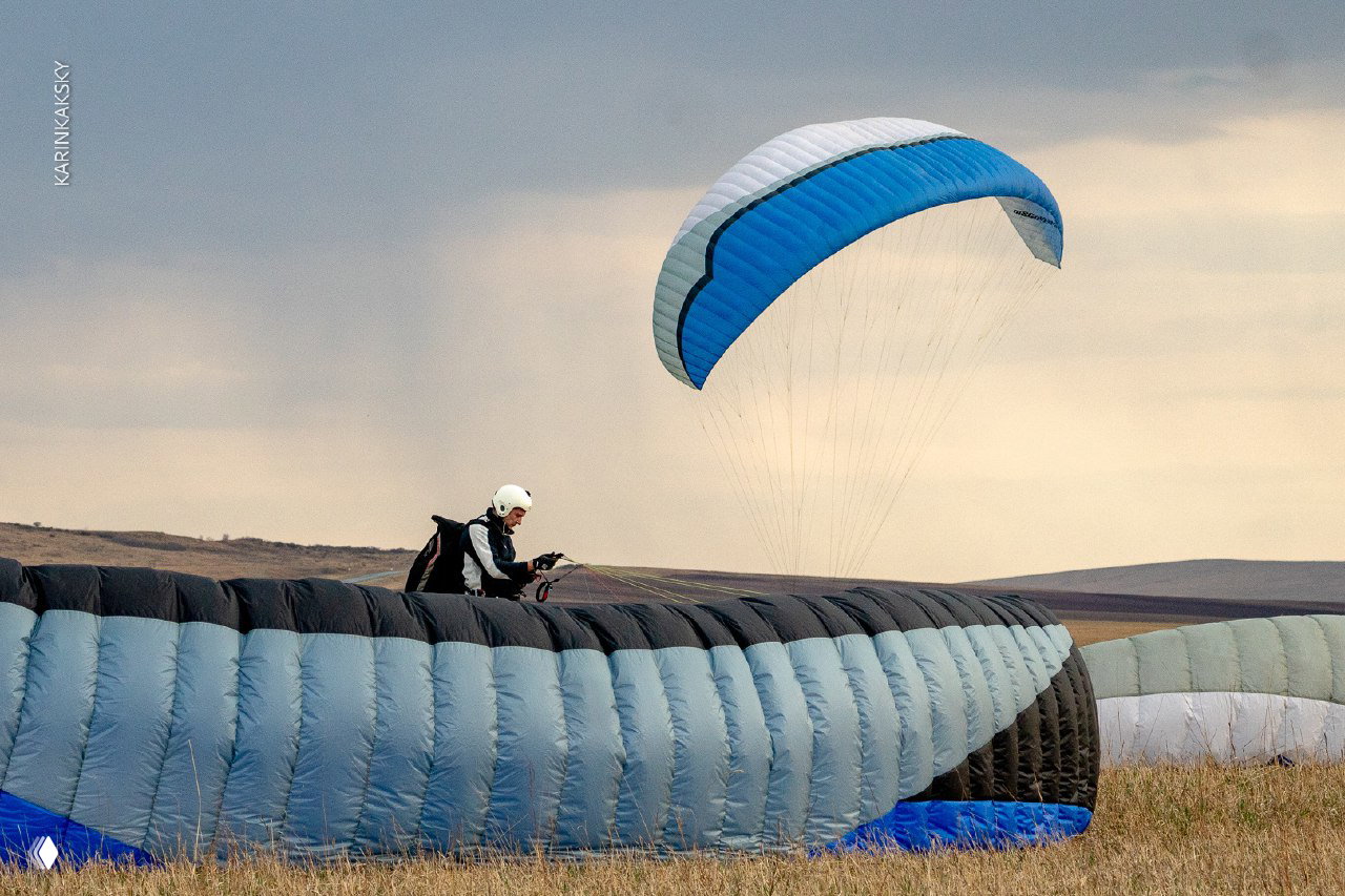 Пилот сидит на земле рядом с длинным рядами уложенных парапланов, над которыми поднимается бело-голубое крыло.