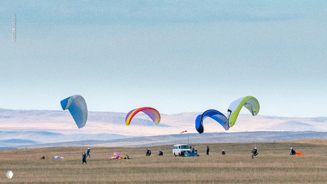 Широкий план: несколько пар пилотов и разноцветных крыльев на большой равнине с автомобилем и дальними холмами.