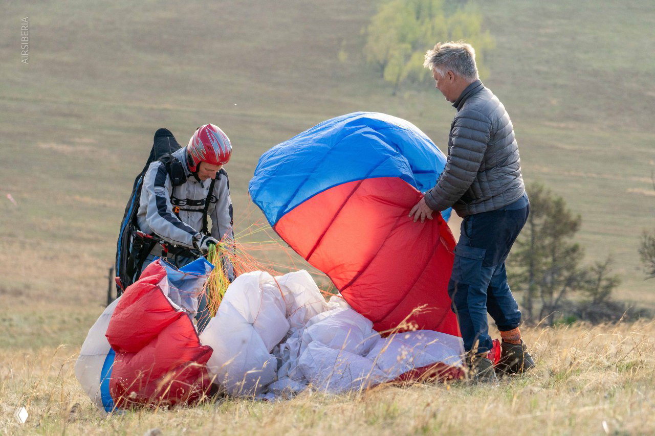 Посадка и уборка купола после полёта: двое человек собирают разноцветный парашютный материал на травянистом склоне.