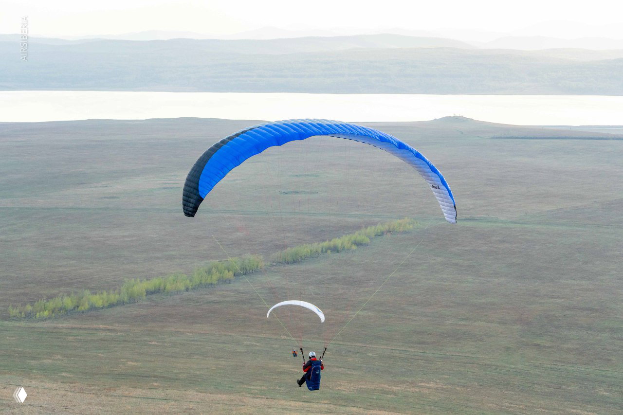 Два параплана в небе над равниной с видимым берегом водохранилища и узкой лесополосой, создающей перспективу маршрута полёта.