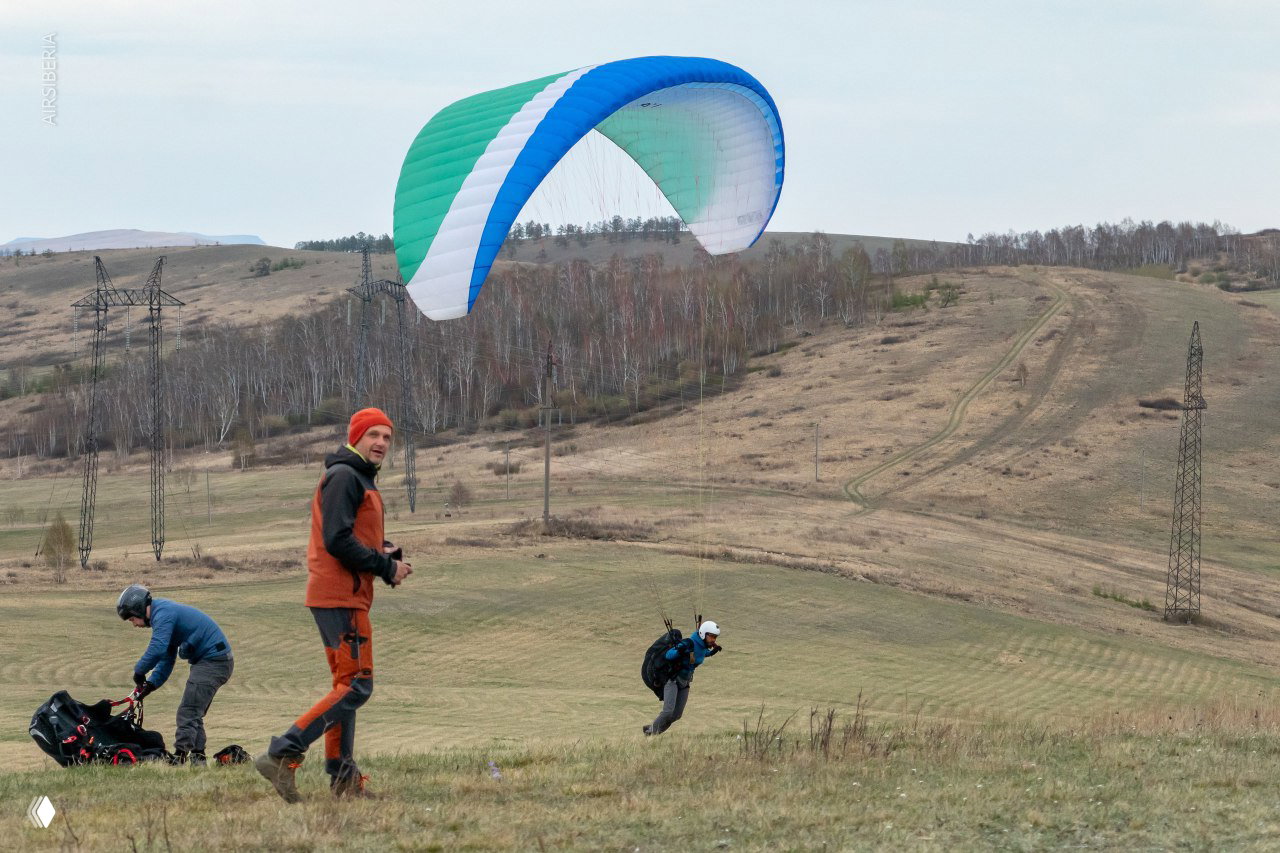 Стартовая площадка: парапланист отталкивается для запуска, рядом другие участники готовят снаряжение на фоне холмов и опор ЛЭП.