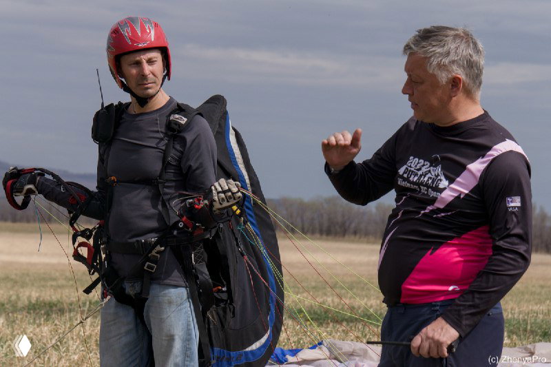 Инструктор разговаривает с пилотом в снаряжении на поле: шлем, подвесная система и стропы видны вблизи подготовки к полёту.