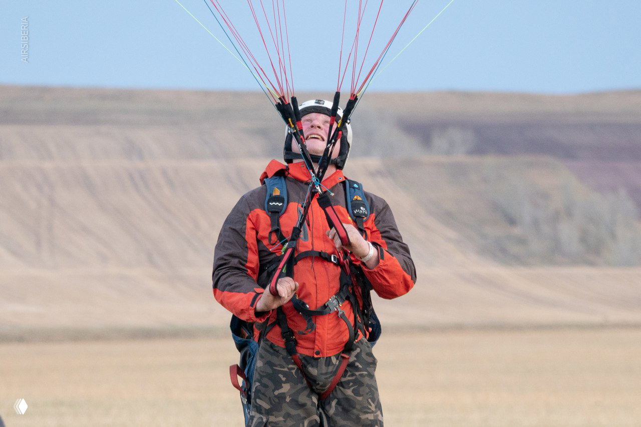 Пилот в красной куртке смотрит вверх и держит стропы параплана во время старта на открытой равнине с холмами на заднем плане.