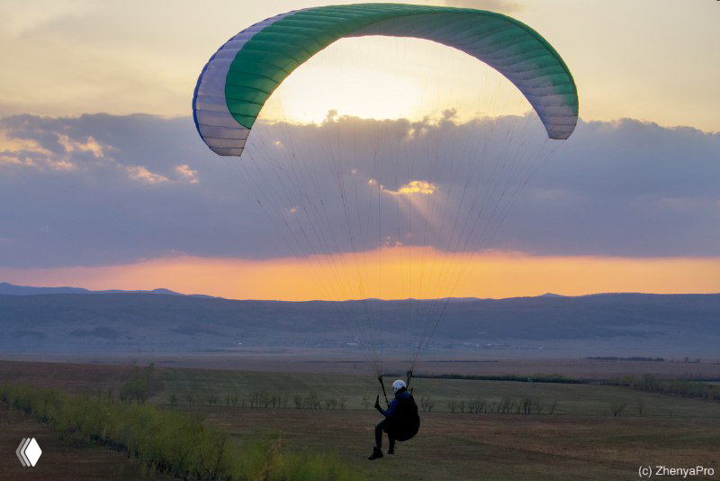 Парапланерист планирует над пейзажем на фоне закатного неба; силуэт купола и горизонт создают ощущение свободы полёта.