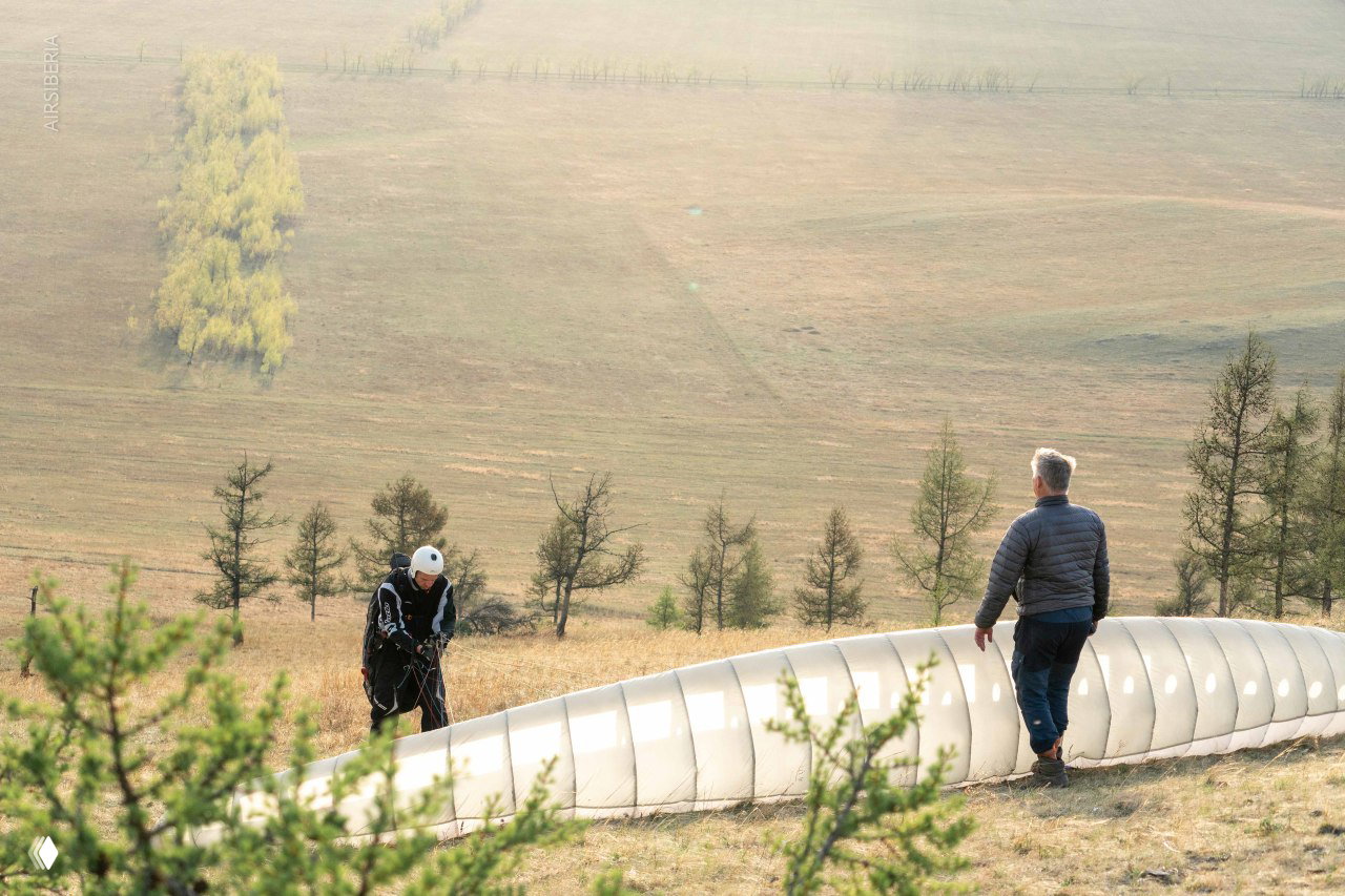 Инструктор и пилот готовят купол параплана на склоне: ткань расправлена, рядом деревья и просторный склон Хакасии перед посадкой.