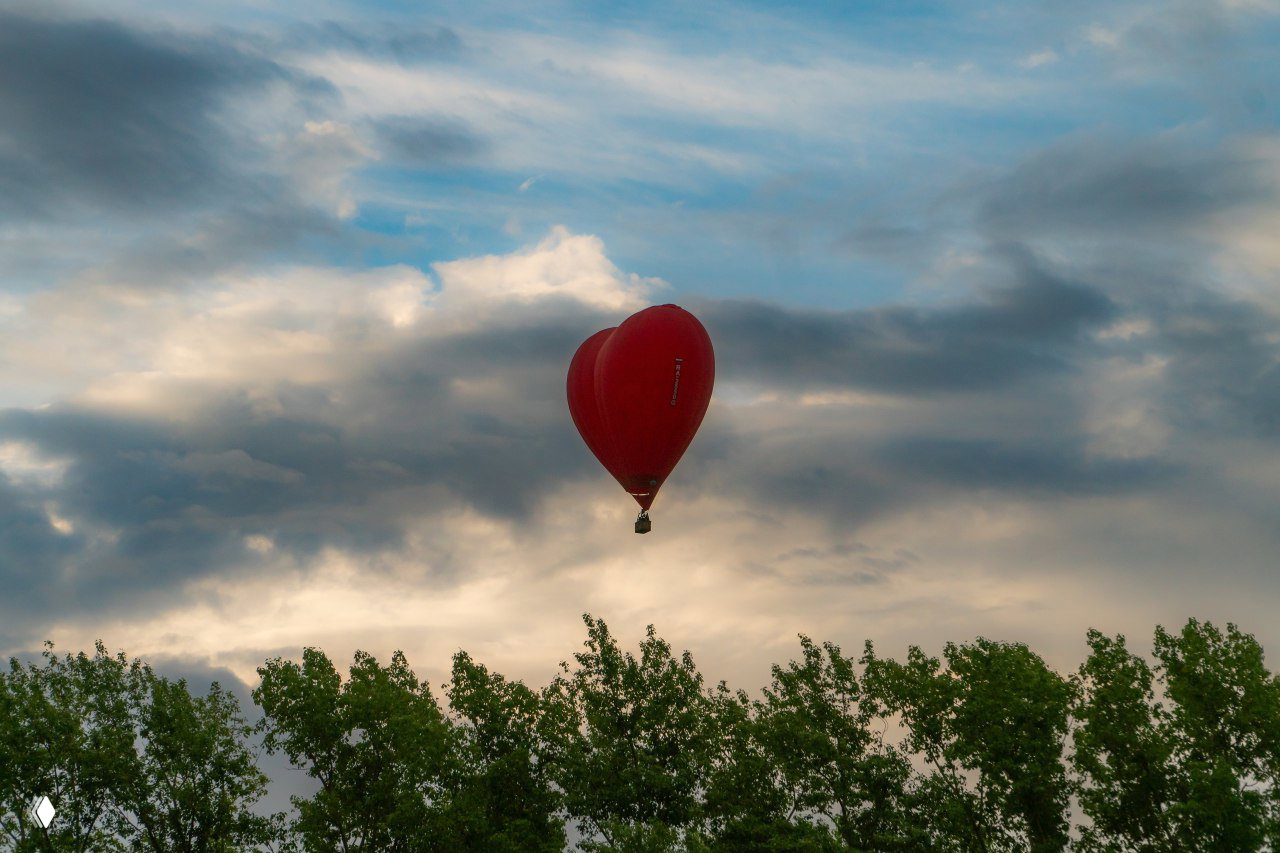 Красный воздушный шар парит над зелёной кромкой леса на фоне драматичного облачного неба — вид с земли и ощущение приближающегося лета.