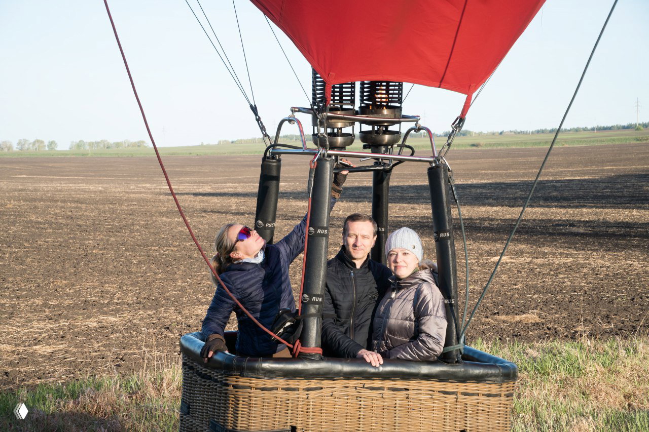 Три человека в корзине воздушного шара на поле; видна конструкция горелки и опорных стоек, один из пассажиров тянется к узлам оборудования перед взлётом.