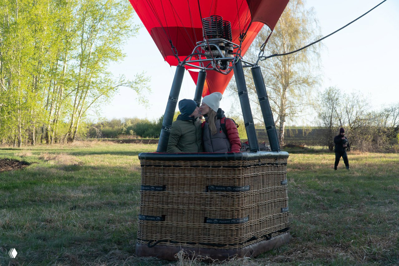 Пара в корзине воздушного шара на фоне молодых лиственных деревьев; они обнялись и целуются у свеже-зелёной кромки леса в мягком свете.