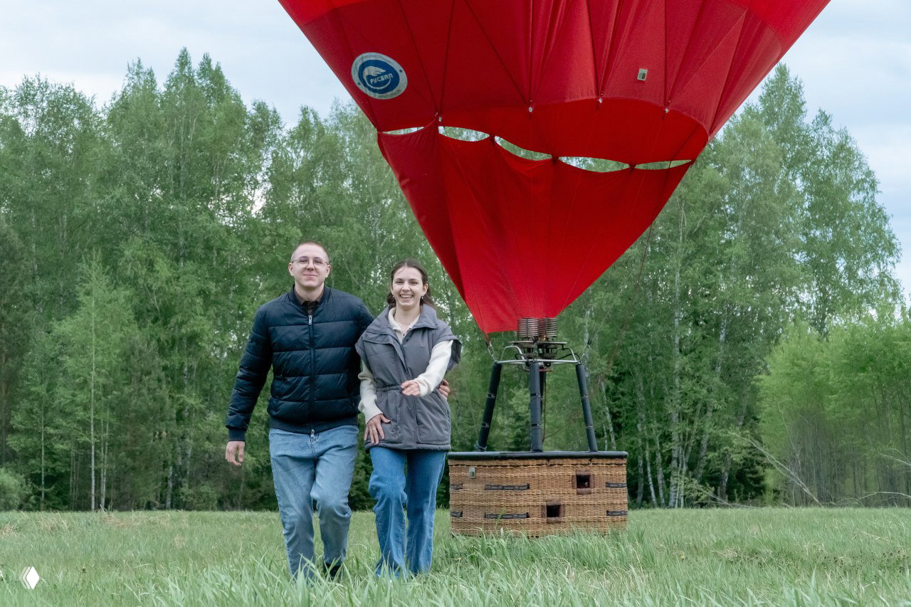 Двое людей идут по травяному полю рядом с опущенным красным воздушным шаром; на заднем плане виден молодый лес и спокойный сельский ландшафт.