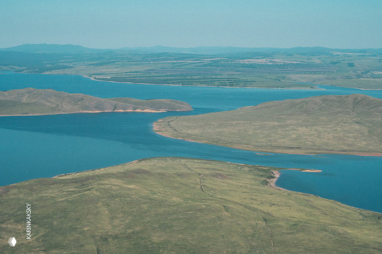 Вид сверху на извилистые берега озера и холмы Хакасии: широкая водная гладь и разреженные сельские участки с дорожками.