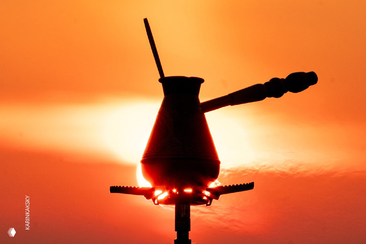 Силуэт кофейной турки на горелке на фоне заходящего солнца; тёплый оранжевый закат создаёт атмосферу привала и кофе с видом.