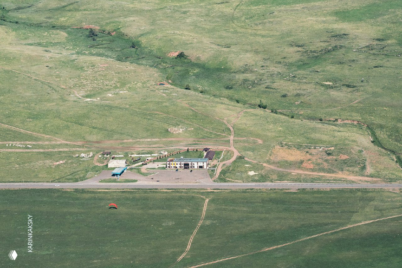 Панорамный вид долины с дорогой и зданием у трассы Красноярск—Абакан; параплан у дороги подчёркивает масштаб местности и маршрут команды.