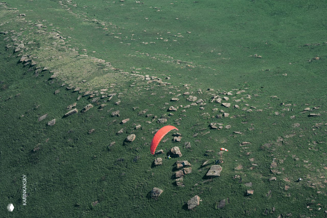 Параплан низко над каменистым участком степи, красное крыло контрастирует с зеленью и рассыпаными валунами, видна посадочная зона рядом с камнями.