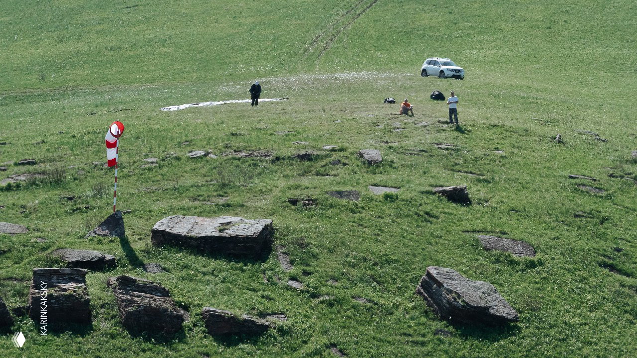 Стартовая площадка с ветровым маркером, людьми и автомобилем на фоне зелёных холмов — подготовка к полётам и проверка ветра перед стартом.