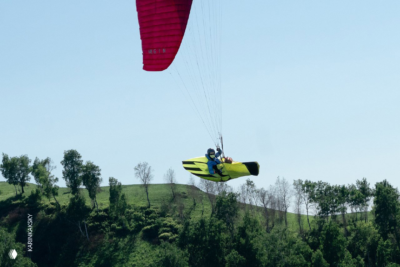 Тандемный полёт с парапланом красно‑жёлтого цвета над лесистыми склонами; пилот и пассажир в подвесной системе видны на фоне неба и деревьев.
