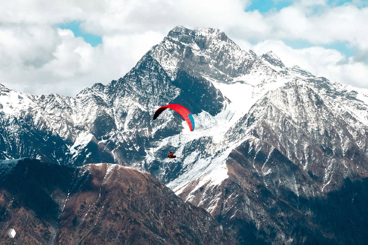 Параплан в небе на фоне заснеженной горной вершины: пилот летит среди предгорий Гималаев, видны рельеф и облака.
