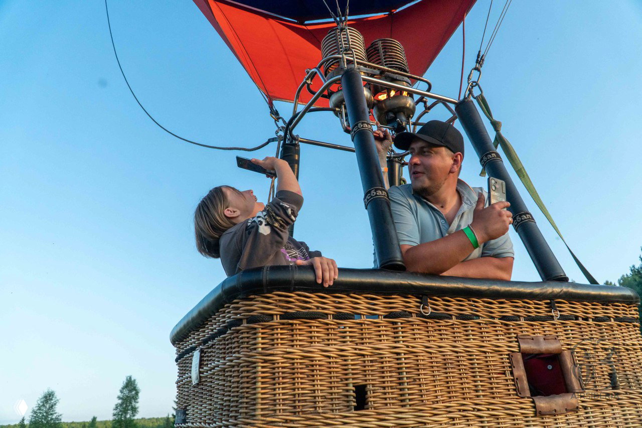 Крупный план корзины воздушного шара с мужчиной и ребёнком рядом с горелкой; дети смотрят вверх, яркое голубое небо на заднем плане.