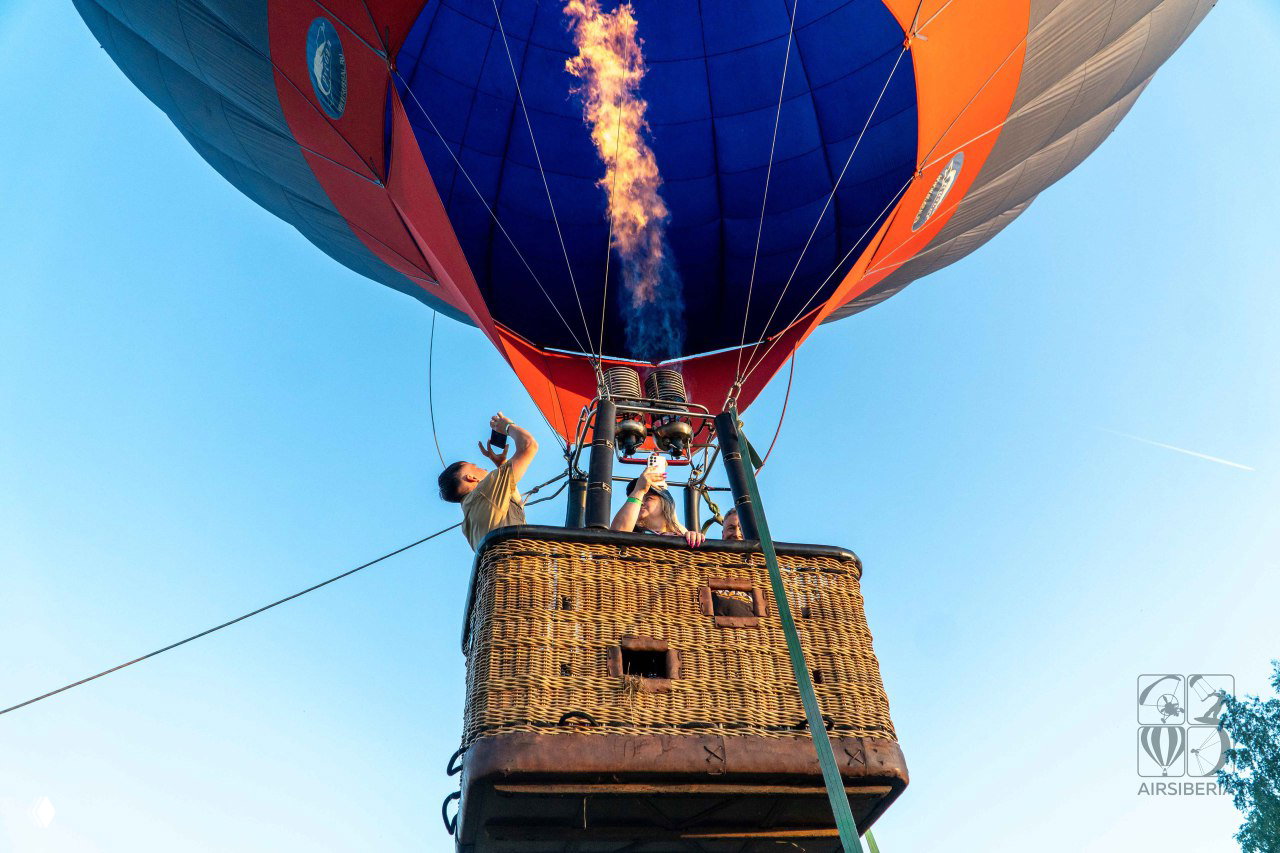 Вид снизу на корзину и включённую горелку: пламя под куполом шара, люди в корзине и трос привязи, небо высоко над головой.