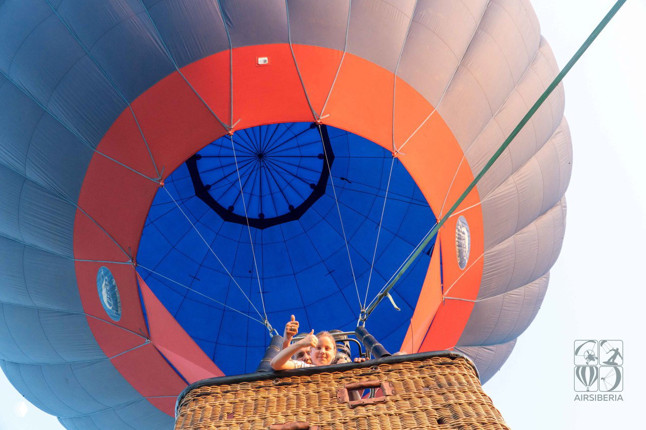 Взгляд снизу вверх на яркий синий и оранжевый купол шара; пассажиры в корзине машут и улыбаются, конструкция и стропы видны крупно.