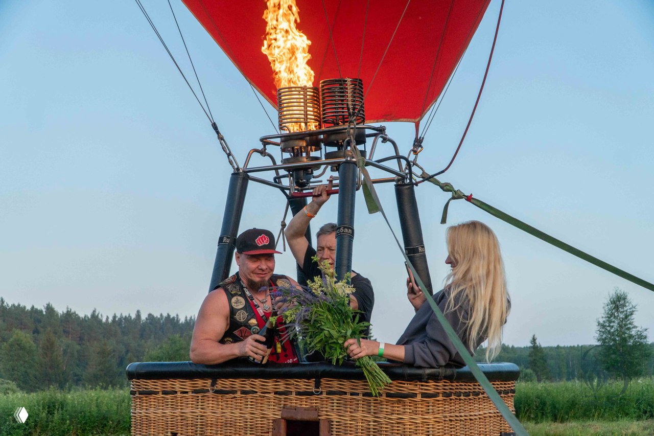 Группа людей в корзине воздушного шара: мужчина с букетом, женщина фотографирует, горелка работает; поле и лес на заднем плане.