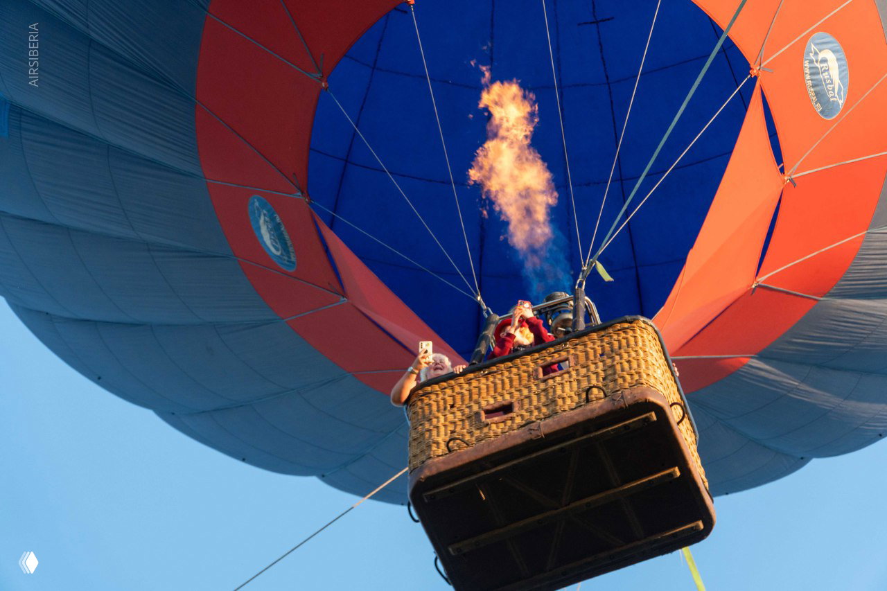 Низкоугловой кадр подъёма шара: корзина с пассажирами, пламя горелки и широкий купол на фоне чистого голубого неба.